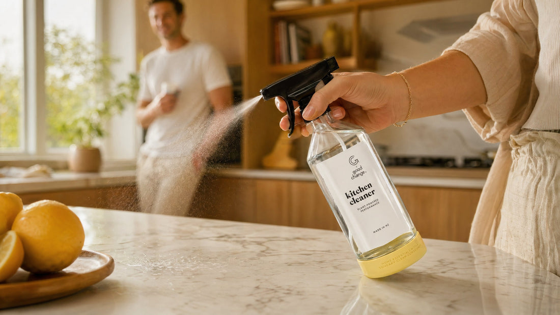 A woman's hand spraying the Good Change Kitchen Cleaner glass bottle onto a marble bench, with lemons in the foreground and a warm Scandi kitchen in the background.