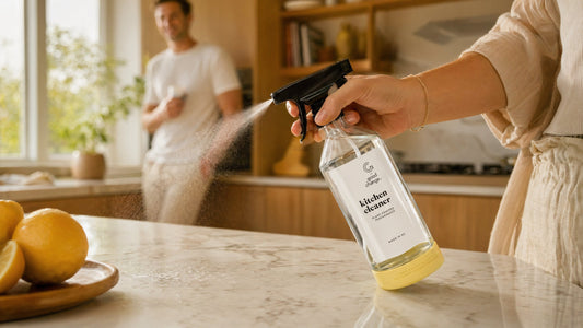 A woman's hand spraying the Good Change Kitchen Cleaner glass bottle onto a marble bench, with lemons in the foreground and a warm Scandi kitchen in the background.