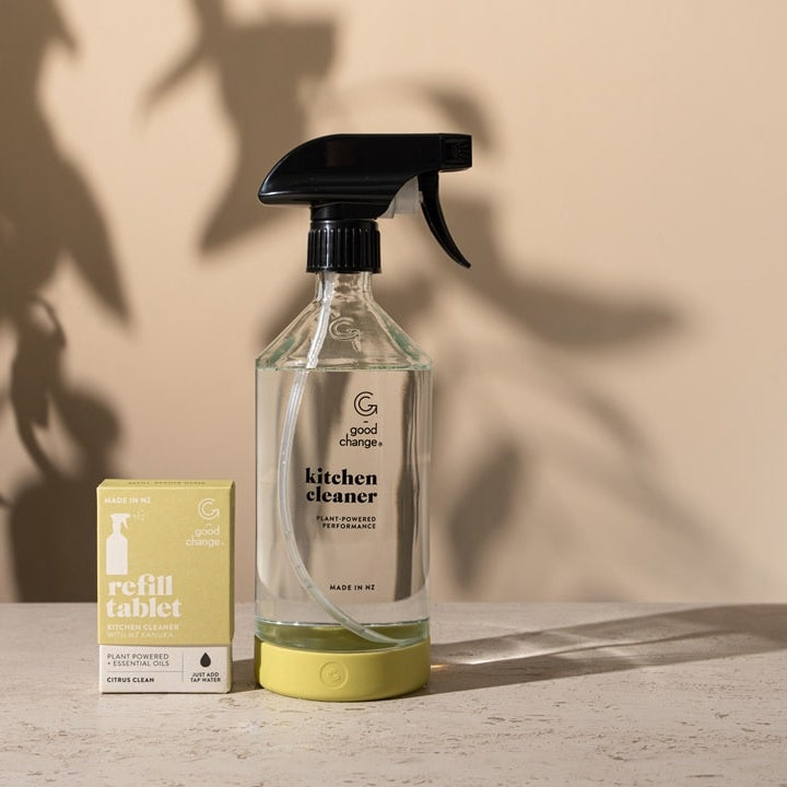 Clear spray bottle labeled 'kitchen cleaner' with a yellow refill tablet on a wooden surface against a beige wall.