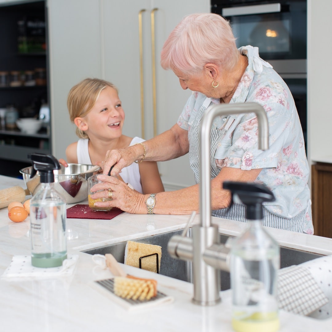A grandmother and granddaughter smile in the kitchen while baking. Good Change's All Purpose Cleaner and Kitchen Cleaner sit proudly on the benchtop.